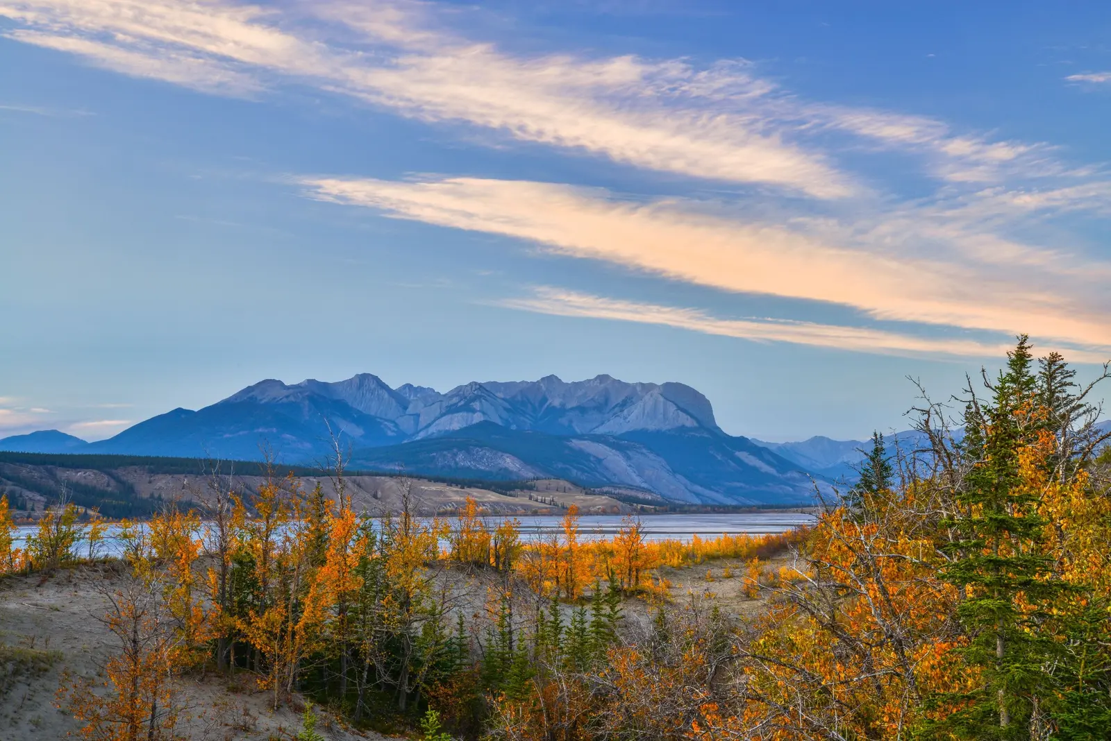 Mountain range above a lake and autumn forest beneath a blue sky.
